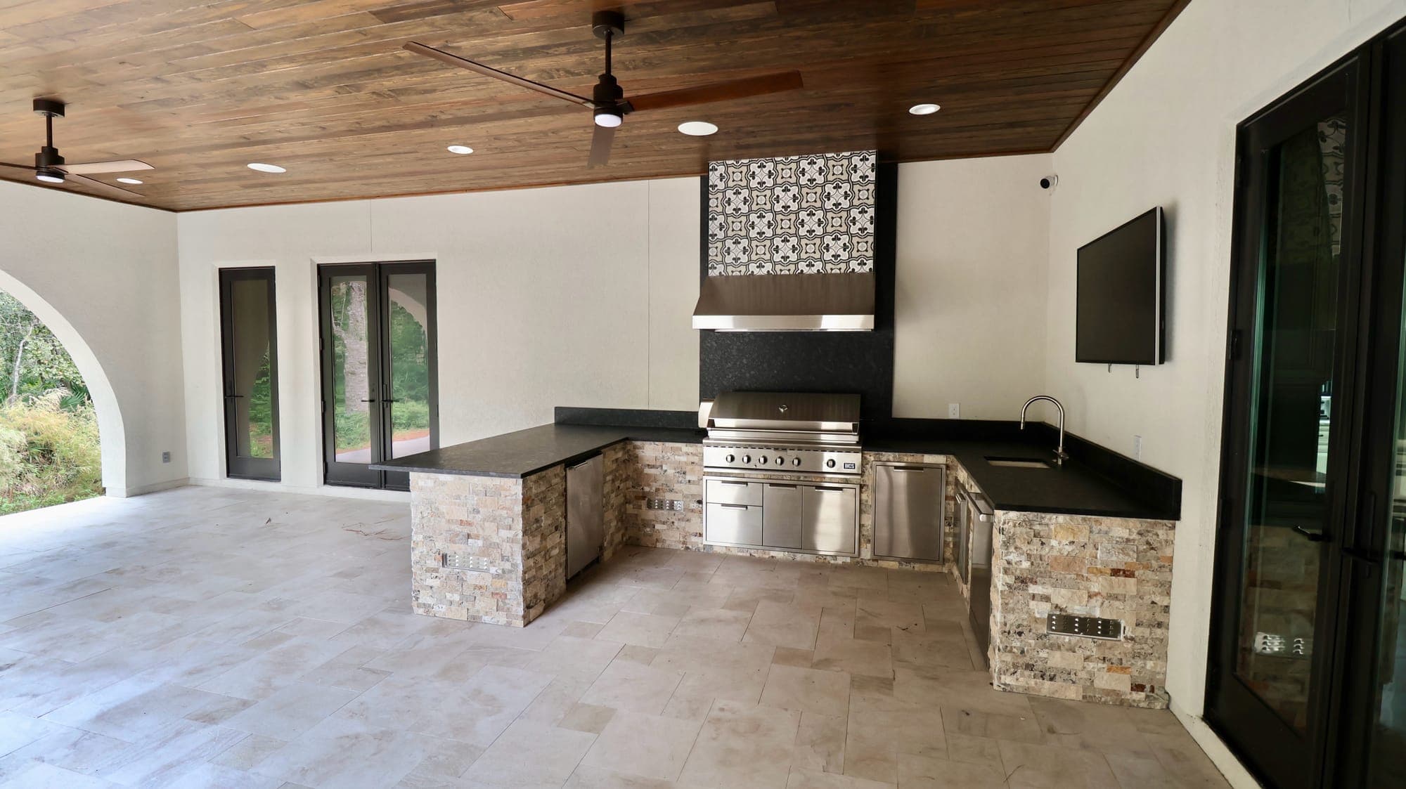 Covered summer kitchen with a stacked-stone island, built-in grill, sink, and tongue-and-groove wood ceiling at a custom home in Nocatee, Florida