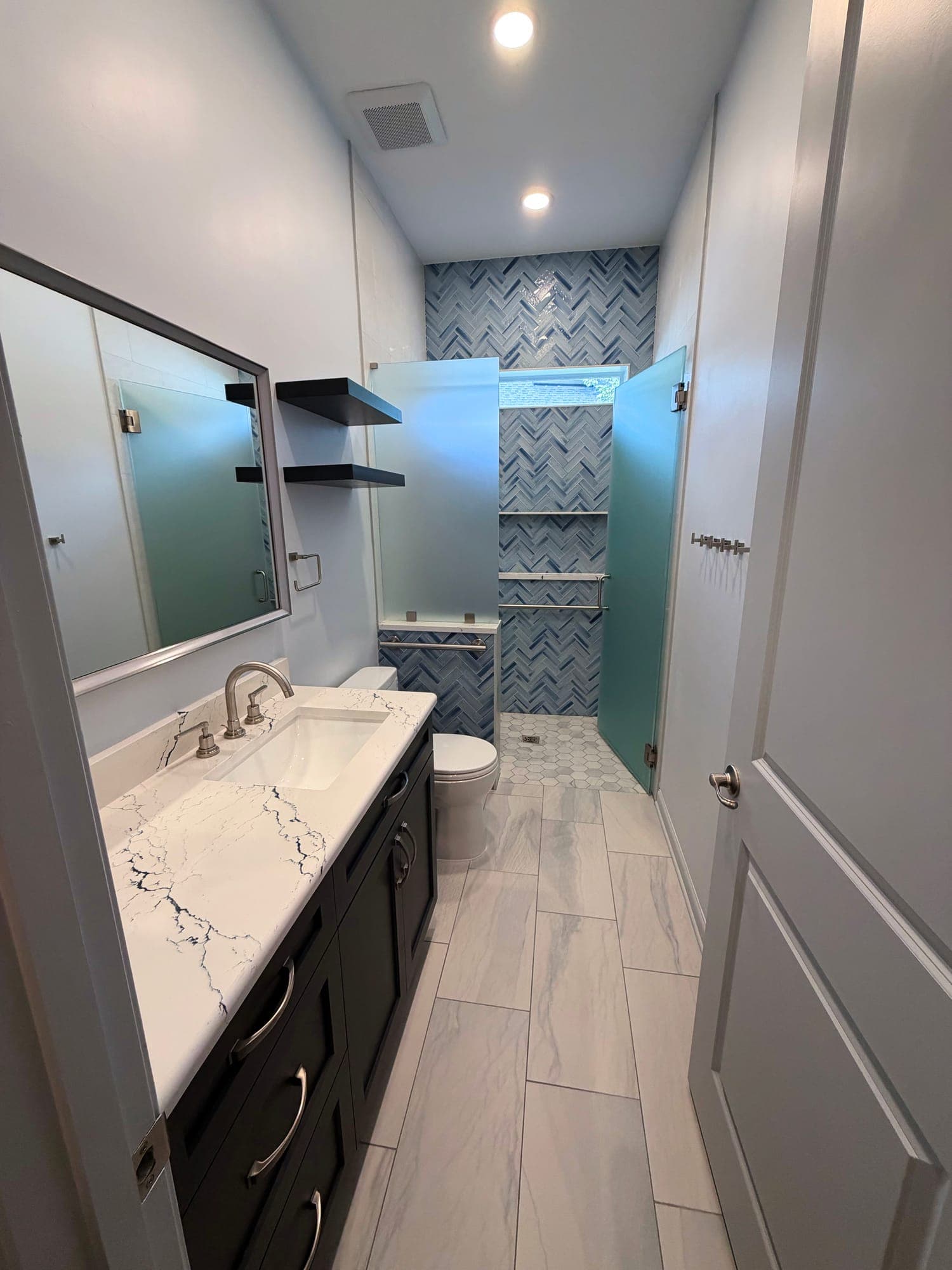 Renovated bathroom with a dark vanity under a white, black-veined countertop, large-format marble-look floor tile, and an open walk-in shower with a blue herringbone tile wall after Integrity Construction Co.'s remodel in Twenty Mile, Nocatee, Florida