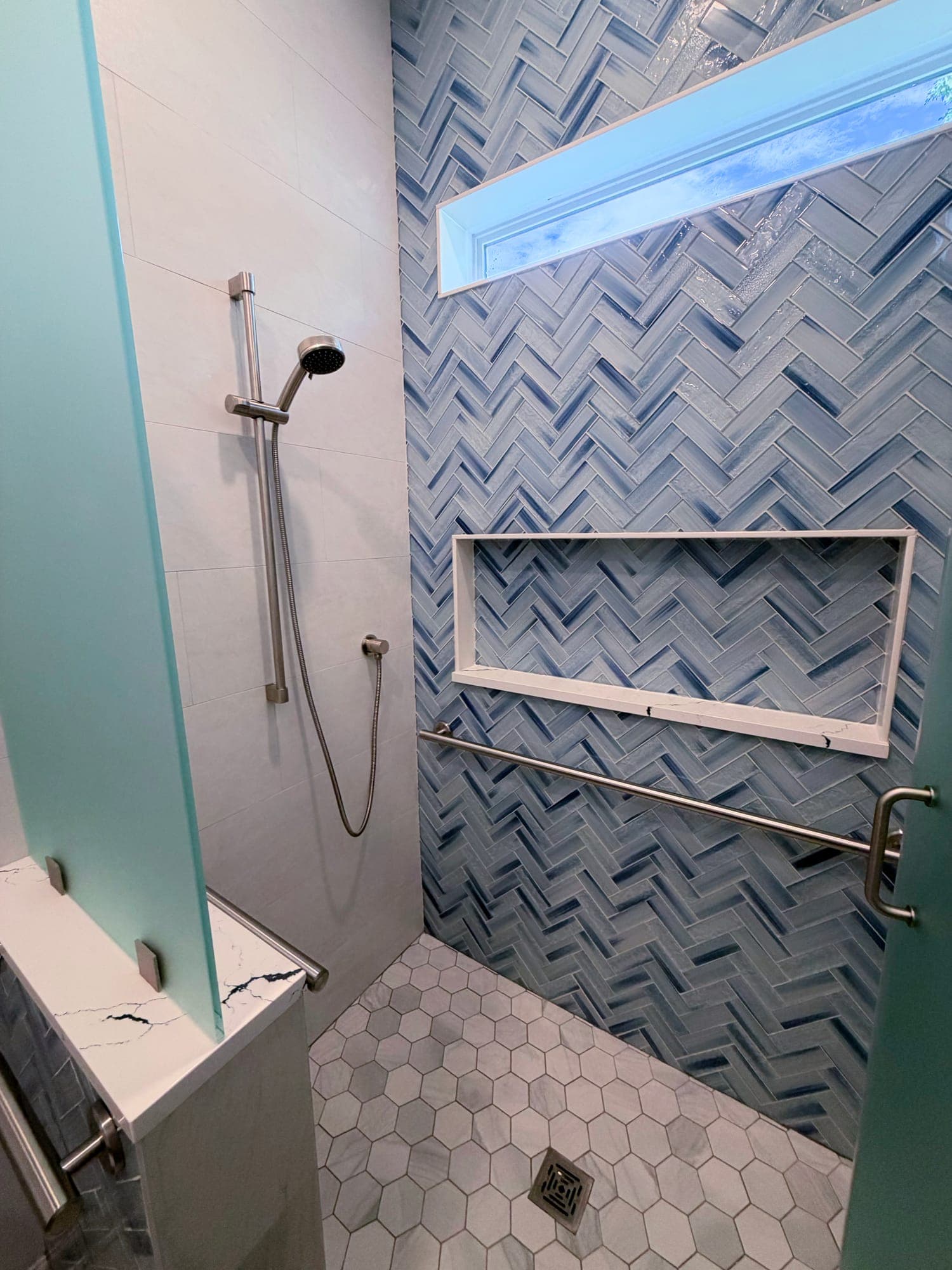 Walk-in shower with a blue herringbone tile feature wall, hexagon mosaic floor, recessed niche, and a handheld shower on a slide bar in a Twenty Mile bathroom remodel, Nocatee, Florida